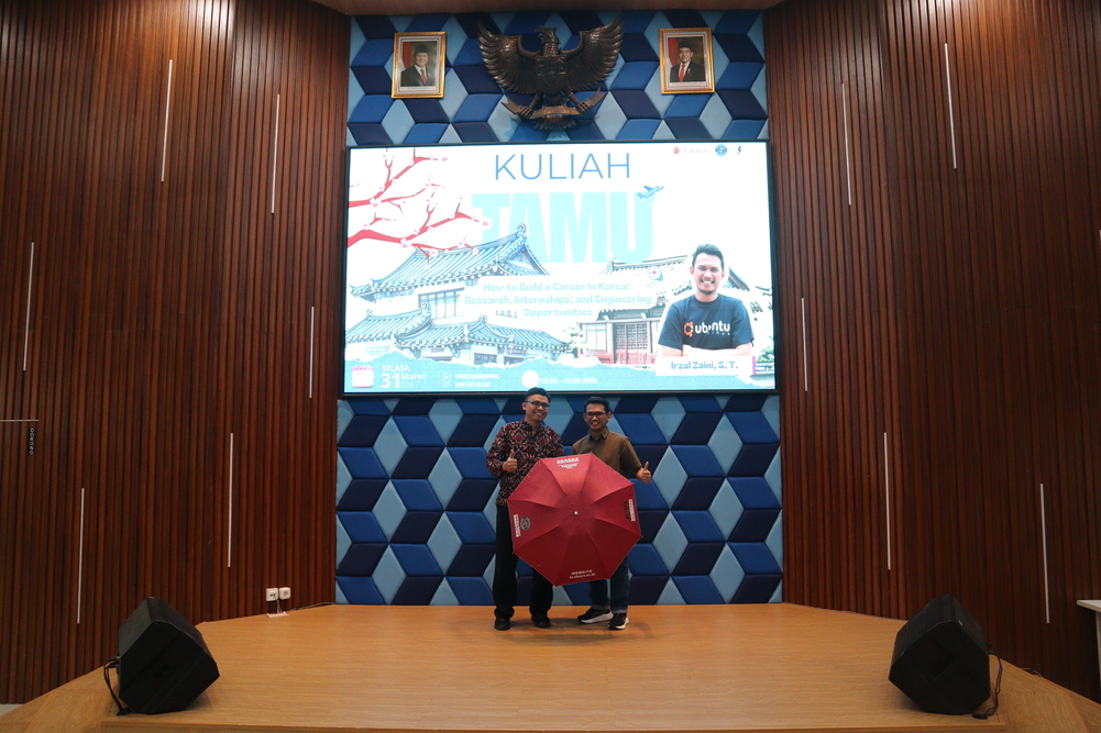 Dua pria berpayung merah di depan slide kuliah tamu Dari Ubaya Sampai Korea